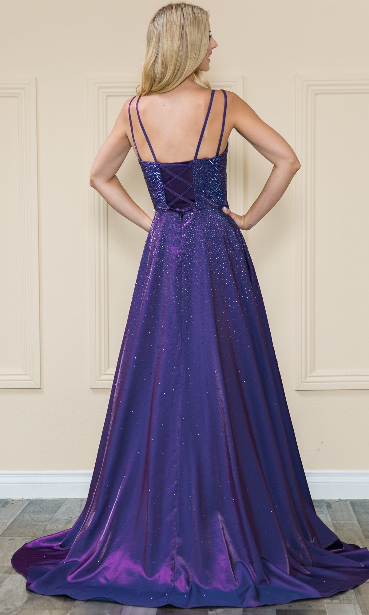 A woman wearing the Long Beaded A-Line Prom Dress: Poly USA 8922, featuring thin straps and a lace-up back, stands facing away from the camera in a floor-length deep purple gown.